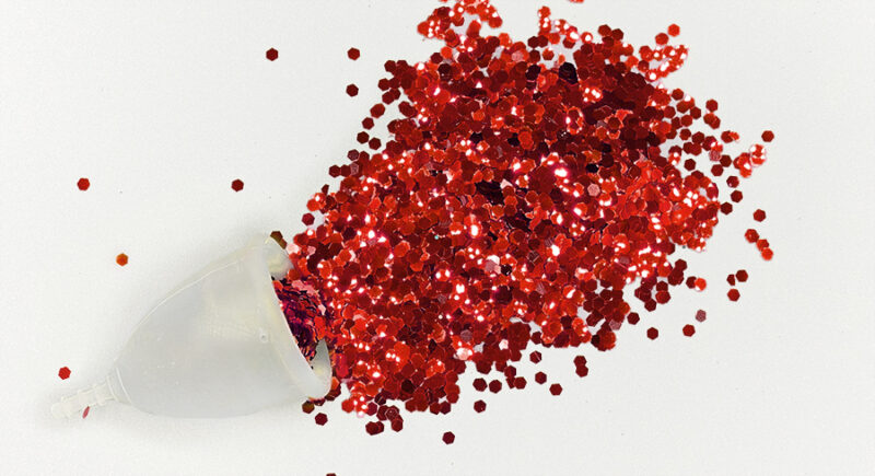 Image of a clear menstrual cup spilling blood red glitter into a puddle.